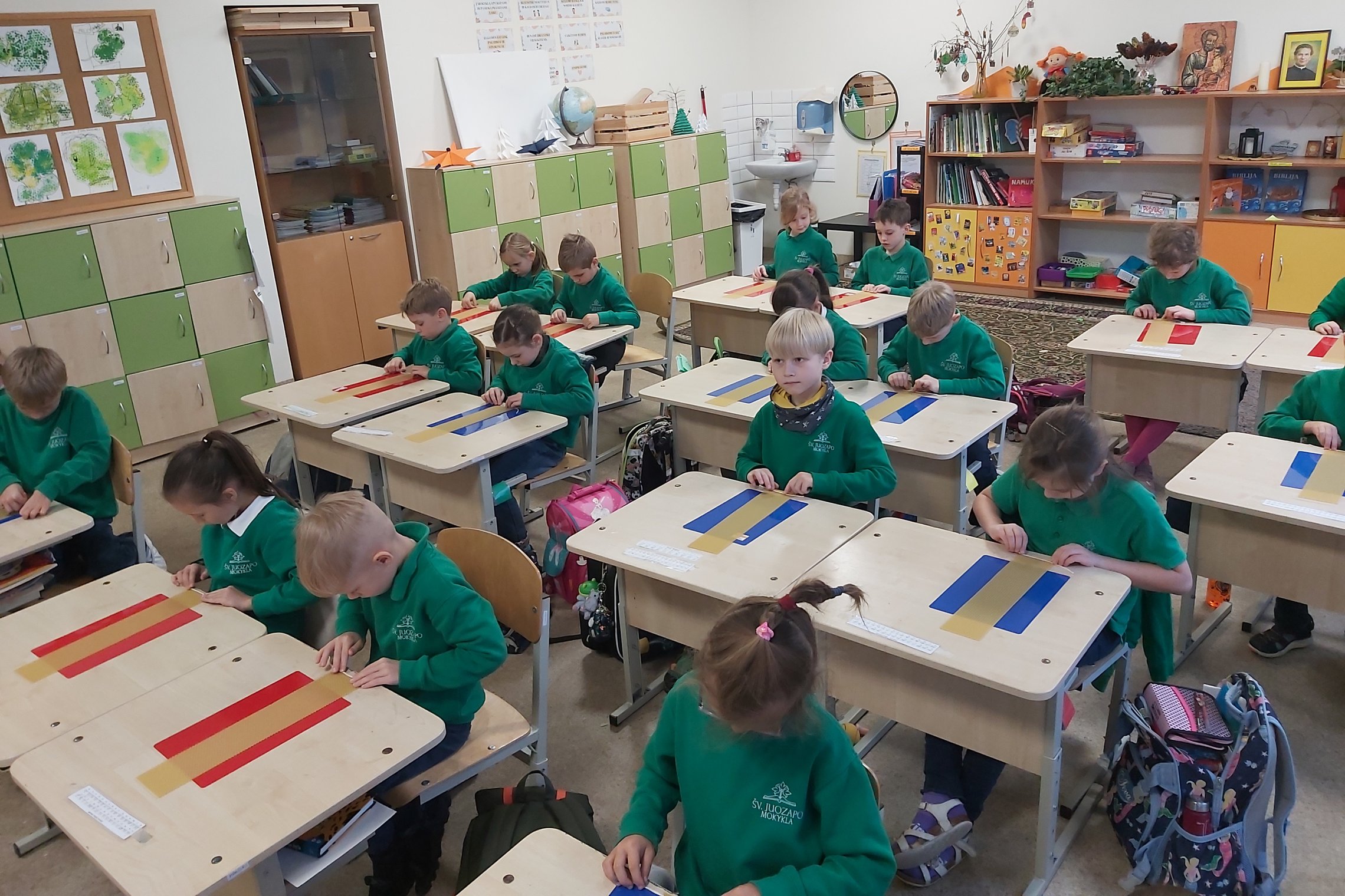 Nuotrauka vaizduoja klasę, joje vaikai sėdi po du, ant stalo padėti spalvoti popieriaus lapai, o ant jų stačiakampiai bičių vaško lakštai. Visi vaikai dėvi tokius pačius megztinius, susikaupę dirba prie lakštų.