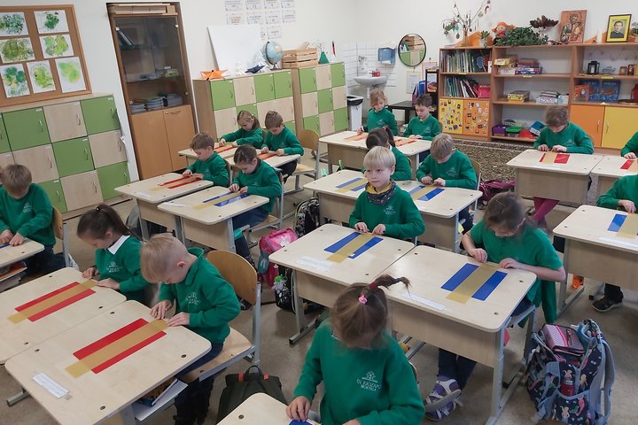 Nuotrauka vaizduoja klasę, joje vaikai sėdi po du, ant stalo padėti spalvoti popieriaus lapai, o ant jų stačiakampiai bičių vaško lakštai. Visi vaikai dėvi tokius pačius megztinius, susikaupę dirba prie lakštų.