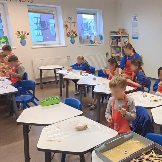 Nuotraukoje yra pavaizduota klasė, kurioje matome daug mažų vaikų. Visi vaikai dėvi raudonas prijuostes, rankose laiko pagaliukus, teptukus arba laiko modelino gabaliukus. Šiais įrankiais jie kuria savo mažiukus projektukus. Beveik visi vaikai sėdi, išsky