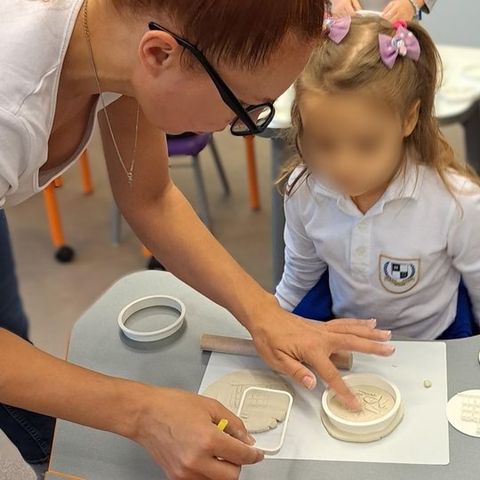 Mokytoja padeda mažai mergaitei kurti molinį atspaudą, naudodama apskritą formelę. Mergaitė dėvi baltus mokyklinius marškinėlius su emblema ir turi dvi rožines kaspino formos plaukų segtukes. Ant stalo – molio plokštelės su įspaustais piešiniais ir kerami