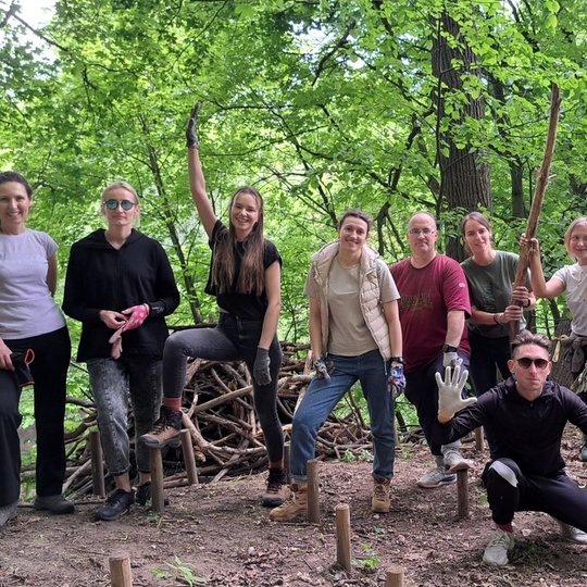 Grupė žmonių stovi miško aplinkoje, kai kurie laiko darbo įrankius ar dėvi pirštines. Fone matyti sukrautos šakos, žemė išmėtyta mediniais kuoleliais – tai gali būti takų, laiptų ar kitos struktūros įrengimo vieta. Visi šypsosi, pozuoja, atmosfera draugiš