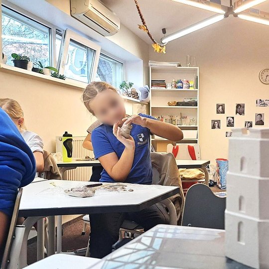 Nuotrauka: vaikai lipdo iš molio kažkokius daiktus. Iš veidų atrodo, kad jie yra susikaupę, stropiai dirba. Veiksmas vyksta uždaroje aplinkoje. Klasėje. Vaikų veidai yra sulieti, nes nuotrauka yra viešinama, tad ne visi vaikai nori rodyti savo veidus.