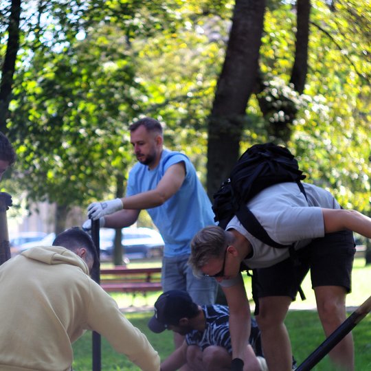 Grupė jaunų vyrų intensyviai dirba parke – jie pritupę ar pasilenkę, įtvirtina medinį stulpą žemėje. Fone matyti žalios vejos, medžių pavėsis ir raudoni suoliukai. Saulėta diena, darbui vyrai pasirinko patogią sportinę aprangą – šortus, marškinėlius, pirš