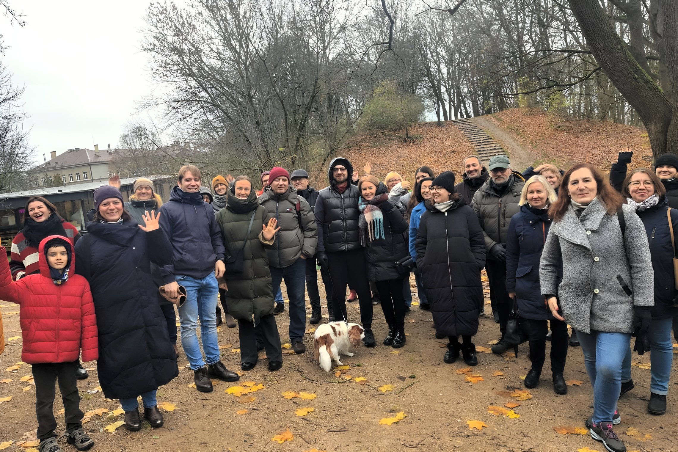 Giedra diena, žmonių grupė pozuoja nuotraukai. Visi apsirengę rudeniškai, su striukėmis, šalikais. Žmonės išsidėstę puslankiu, viduryje stovi mažas rudai baltas šuo. Fone matomas nedidelis kalniukas, ant jo augantys medžiai jau numetę lapus.