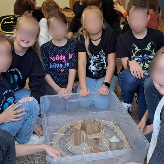 Grupė vaikų sėdi aplink plastikinę dėžę, kurioje sukurtas pilies maketas iš kartono, medienos ir smėlio. Makete matyti gynybinė siena, bokštai, vartai ir pastatai. Vaikai šypsosi, vilki įvairius marškinėlius su piešiniais ir užrašais. Fone – klasės aplink