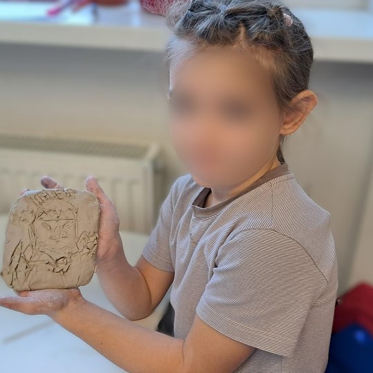 Vaikas su dryžuotais marškinėliais rankose laiko molio plokštelę, kurioje išraižytas žmogaus veidas – tikėtina, kunigaikštis ar istorinis personažas. Fone matyti klasės interjeras: stalas, radiatorius ir palangė su daiktais.