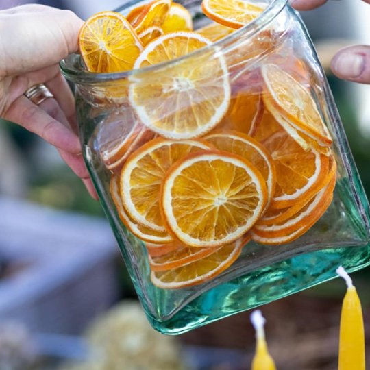 Nuotraukoje stambiu planu pavaizduotas stačiakampinis stiklainis, kuris yra pilnas apelsinų skiltelių. Stiklainį laiko dvi raukšlėtos rankos. Taip pat nuotraukoje neryškiai matosi dvi geltonos vaškinės žvakės.