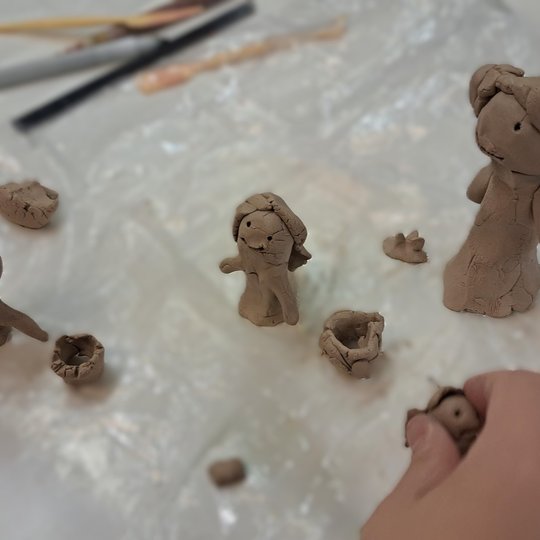 Nuotraukoje vaizduojamas stalas, apdengtas plastmase, fone ant stalo guli keli įrankiai moliui. Nuotraukos apačioje kadre pasirodo ranka, laikanti molio gabaliuką, o ant plastmasės taip pat padėti keli moliniai žmogeliukai.