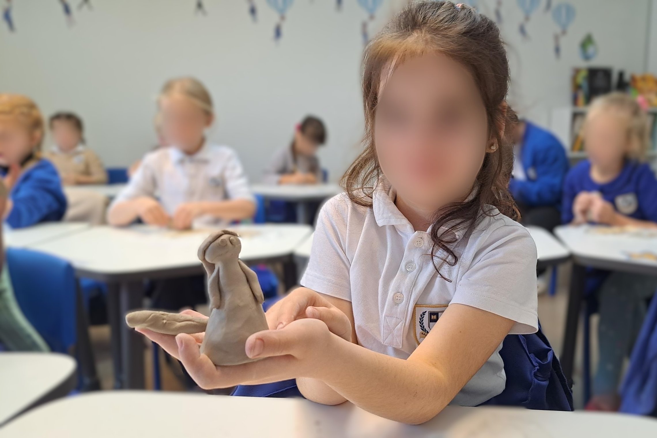 Pirmajame nuotaukos plane matome mergaitę, kuri ištiesusi rankas laiko iš molio nulipdytą moters figūrėlę. Fone matome kitus vaikus, atsisėdusius už mokyklinių stalų, kurie irgi lipdo molines figūrėles. Vaikai apsirengę mėlynomis ir baltomis uniformis.