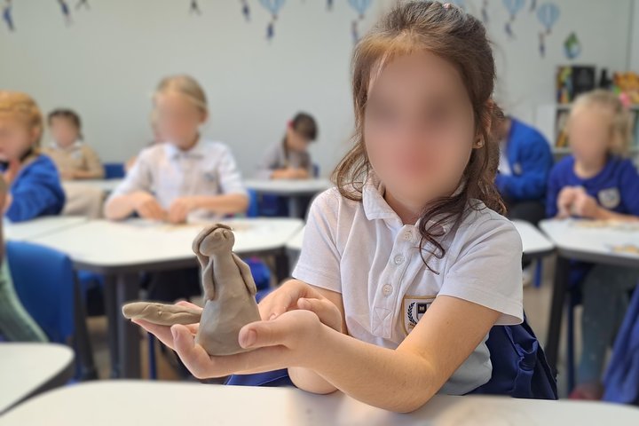 Pirmajame nuotaukos plane matome mergaitę, kuri ištiesusi rankas laiko iš molio nulipdytą moters figūrėlę. Fone matome kitus vaikus, atsisėdusius už mokyklinių stalų, kurie irgi lipdo molines figūrėles. Vaikai apsirengę mėlynomis ir baltomis uniformis.