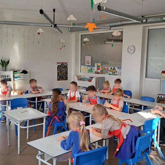 Nuotraukoje matome dvylika klasėje sėdinčių vaikų ir vieną auklėtoją. Visi vaikai dėvi raudonas prijuostes ir lipdo Gedimino bokštą iš moliu. Priešais vaikus ant stalo pastatytas Gedimino bokšto figurėlė. Klasėje galime matyti lubose kabančius debesėlius 