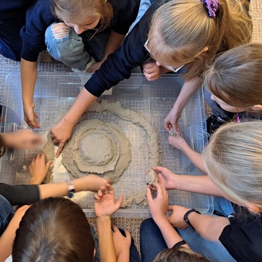Nuotraukoje vaikai pritūpę aplink plastikinę dėžutę, kurioje konstruoja būsimą piliakalnį, daro iš kinetinio smėlio apskritimą. Galima įžvelgti piliakalnio įdubimus, gynybinę sieną.