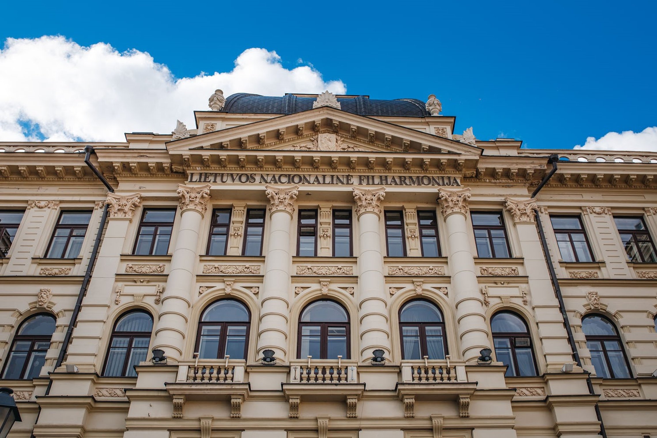 Istoristinio stiliaus pastatas su kolonomis ir puošybinėmis detalėmis, viršuje matomas užrašas „Lietuvos nacionalinė filharmonija“. Pastato fasadas šviesus, langai tamsiai rudi, viršuje – kupolinio stogo fragmentas. Fone – mėlynas dangus su baltais debesi