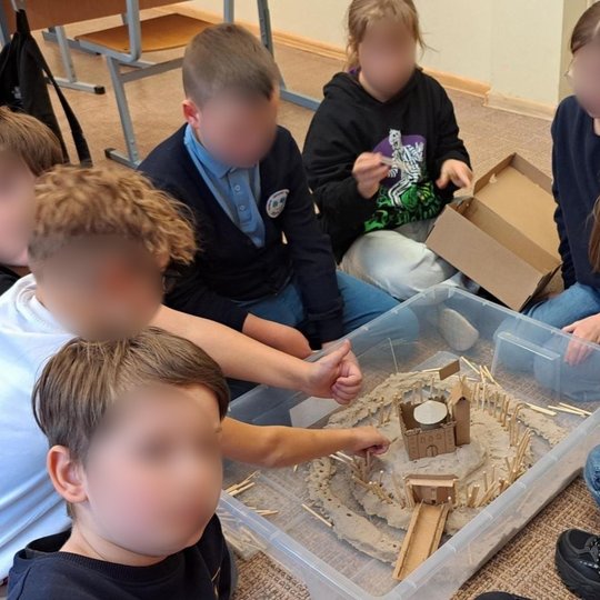 Nuotraukoje vaikai šypsosi ir džiaugiasi savo rankomis pastatytu piliakalniu. Mokiniai labai detaliai atvaizdavo senovės pilį ant piliakalnio - iš kartono pastatė dviaukštę pilį su bokštais, mediniais pagaliukais pavaizdavo ramsčius, prilaikančius konstru