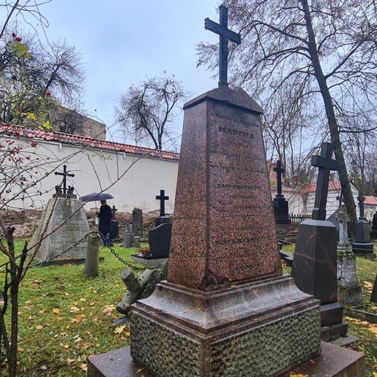 Nuotraukos priekiniame plane matomas stambus akmeninis antkapis. Antkapis ganėtinai aukštas ir smailėjantis, jo viršūnėje pritaisytas metalinis kryžius. Nuotaukos fone matomi kiti panašių gabaritų antkapiai ir juodai apsirengęs žmogus einantis palei kapin
