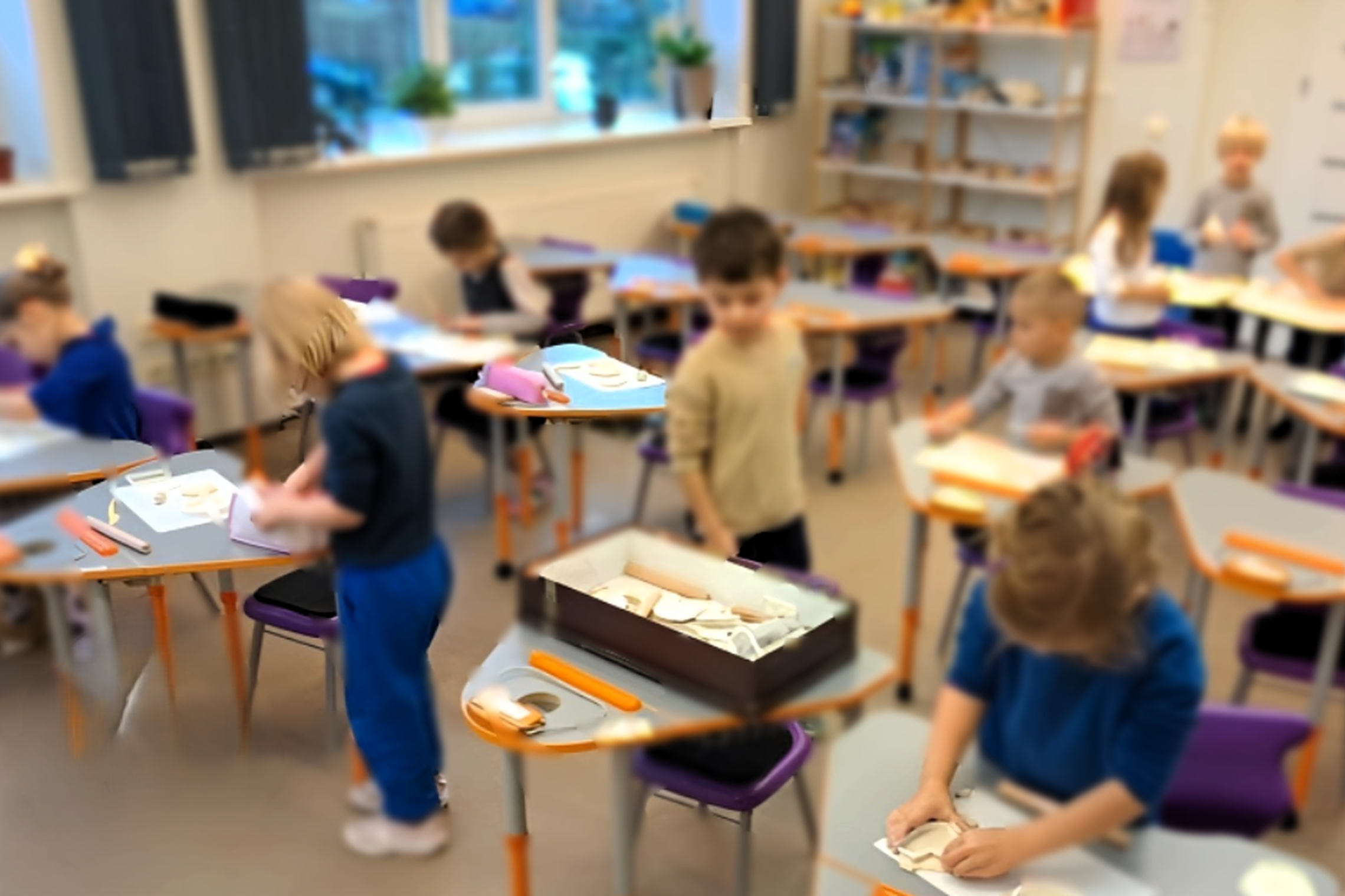 Nuotrauka daryta mokyklos klasėje. Matome aštuonis vaikus, kurie dėvi mėlynas mokyklines uniformas. Kai kurie vaikai stoviniuoja, kiti iš molio lipdo įvairias figūras. Nuotrauka yra išsiliejusi, todėl konkrečios detalės nėra matomos