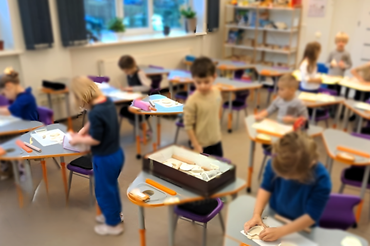Nuotrauka daryta mokyklos klasėje. Matome aštuonis vaikus, kurie dėvi mėlynas mokyklines uniformas. Kai kurie vaikai stoviniuoja, kiti iš molio lipdo įvairias figūras. Nuotrauka yra išsiliejusi, todėl konkrečios detalės nėra matomos