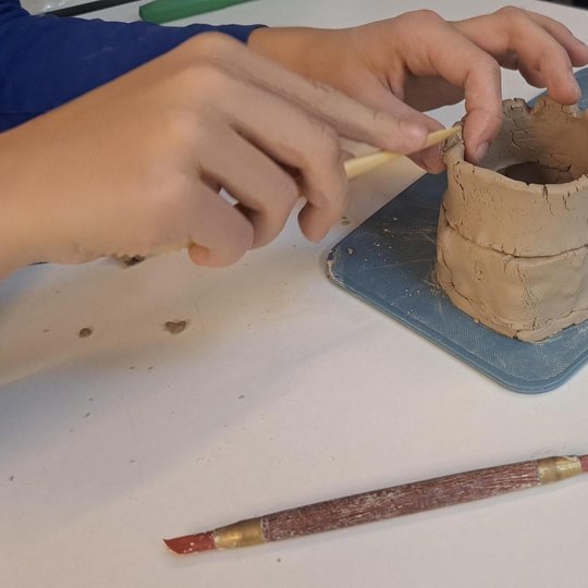 Nuotrauka: vaiko rankos lipdo kažką iš molio ant plastikinės lentelės. Šalia yra padėti įrankiai. Pagaliukas ir metalinė mentelė. Veiksmas vyksta klasėje. Pirštai labai atsakingai dirba su lipdiniu.