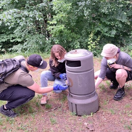 Trys žmonės susikaupę valo ar perdažo pilką lauko šiukšliadėžę gamtoje. Visi dėvi pirštines, du iš jų – respiratorius, vienas turi kuprinę. Šalia matyti valymo priemonių, purškiamų dažų flakonai, pirštinės ir šluostės. Fone – tanki žaluma, medžių lapija.