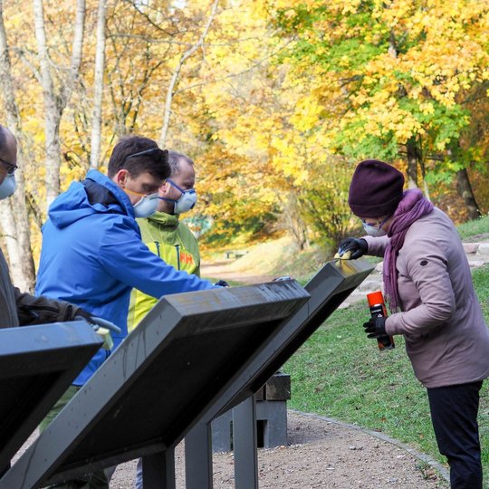 Keli žmonės dėvi apsaugines kaukes ir pirštines, dirba lauke rudens fone. Jie valo arba atnaujina informacinius stendus, greičiausiai parko ar kultūrinio objekto teritorijoje. Viena moteris naudoja purškiamą priemonę ir šluostę, kiti taip pat susitelkę į