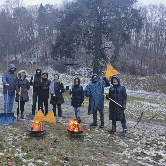 Nuotraukoje vaizduojama aštuonių žmonių grupė, kuri stovi ant žolės netoli kelio. Fone yra nedidelis kalnas, apaugęs medžių. Kai kuriose aplinkos vietose yra iškritusio sniego. Visi žmonės yra apsirengę šiltai su striukėmis. Dauguma jų rankose laiko grėbl