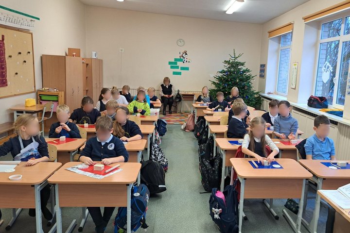 Nuotraukoje klasėje sėdintys nedideli vaikai. Suolai išdėlioti dviem eilėmis, už jų klasės gale sėdi moteris. Prie galinės sienos stovi papuošta kalėdinė eglutė. Prieš kiekvieną vaiką ant stalo padėta lentelė su lipdymui skirto molio gabalėliu, pagaliukas