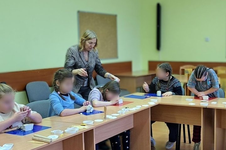 Nuotrauka: veiksmas vyksta uždaroje aplinkoje, kurioje šeši vaikai lipdo iš molio darbelius. Aplinkui pridėta daug visokių, lipdymui skirtų formelių. Prie vaikų stovi viena moteris, padedanti vaikams gražiai padaryti lipdinukus iš molio.