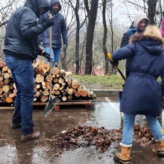 Nuotraukoje nedidelė grupė žmonių tvarko aplinką. Du iš jų priekyje grėbia lapus ant šlapio, asfaltuoto kelio, o kiti grupės nariai stovi gale ir stebi. Fone, ant kelio stovi medinis padėklas su tvarkingai sukrautomis malkomis. Gale augantys medžiai yra n