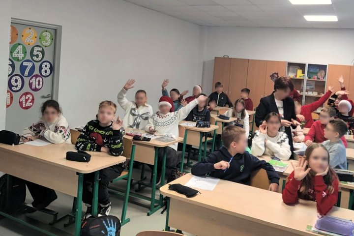 Nuotrauka yra nufotografuota klasėje, kurioje prie suolų sėdi dvidešimt vaikų ir stovi auklėtoja. Trylika vaikų žiūri į kamerą ir mojuoja rankomis, o kiti klausosi ką aiškina auklėtoja. Keli vaikai ant savo galvų yra užsidėję raudona Kalėdų senelio kepurę