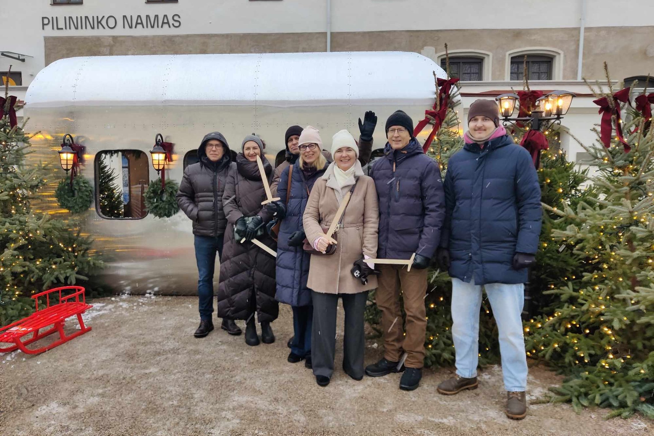 Nuotraukoje septynių žmonių grupė stovi prieš sidabrinį vagonėlį, papuoštą žibintais ir vainikais. Fone yra pastatas, ant kurio didelėmis raidėmis užrašyta „Pilininko namas“. Už žmonių yra trys puoštos eglutės, taip pat kitoje pusėje stovi dar viena eglut