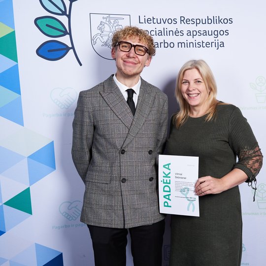 Nuotraukoje prie Lietuvos Respublikos socialinės apsaugos ir darbo ministerijos foto sienos pozuoja du žmonės. Kairėje pusėje vyras su pilku švarku, šviesiais garbanotais plaukais ir akinukais, dešinėje šviesiaplaukė moteris tamsiai žalia suknele, belaika