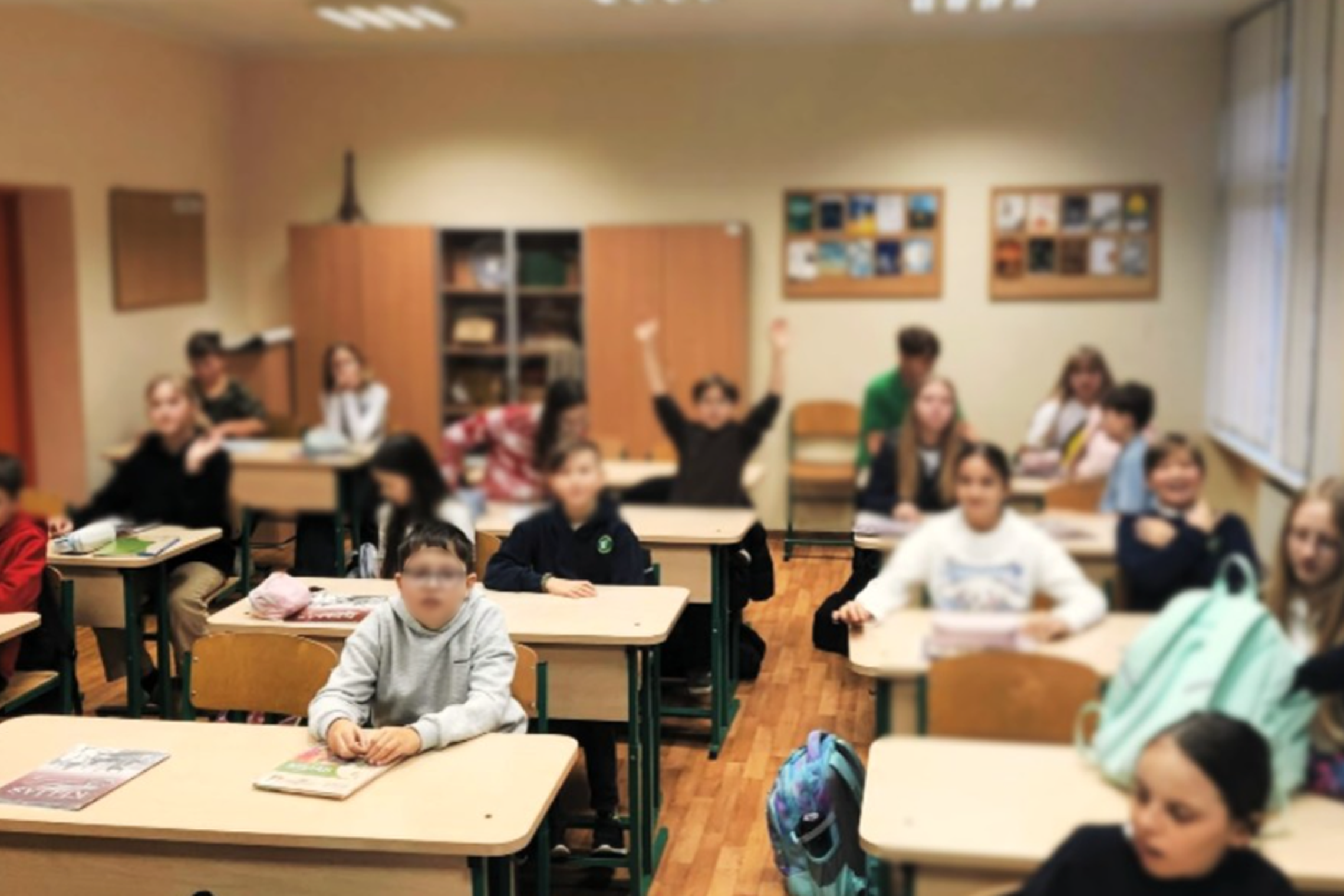 Klasėje sėdi mokiniai prie savo suolų, kai kurie žiūri į priekį, kiti kalbasi tarpusavyje. Vienas berniukas klasės gale pakėlęs rankas į viršų. Priekiniai stalai padengti vadovėliais ir kuprinėmis. Matoma šilta, šviesi klasės aplinka su medinėmis grindimi