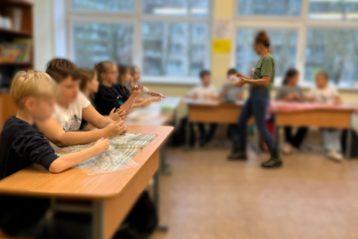 Nuotrauka: veiksmas vyksta uždaroje aplinkoje, panašu, kad mokykloje. Aplink stalus yra susėdę daug vaikų ir per centrą stovi jauna moteris, pasakojanti kažką vaikams, o vaikai jos įdėmiai klausosi.
