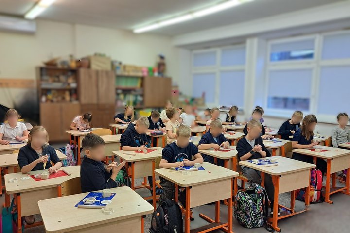 Nuotraukoje vaizduojama nemaža mokyklinė klasė. Joje prie suolų sėdi mokiniai, kurie iš balto molio lipdo įvairias figūras. Pro klasės langus matosi, kad lauke sutemę. Galima manyti, kad nuotauka daryta anskti ryte. Vaikai dėvi tamsiai mėlynas mokyklines