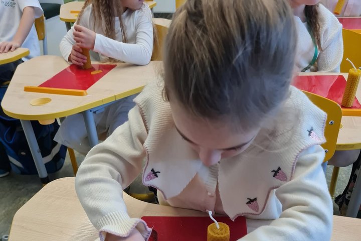 Nuotraukos pirmame plane yra matoma mokyklinio amžiaus mergaitė, kuri iš vaško metalinėmis formelėmis pjausto įvairias figūras. Šalia figūrų stovi vaškinė žvakė, ant kurios figūros turbūt bus klijuojamos. Mergaitė apsirengusi smėlio spalvos megztiniu. Ant
