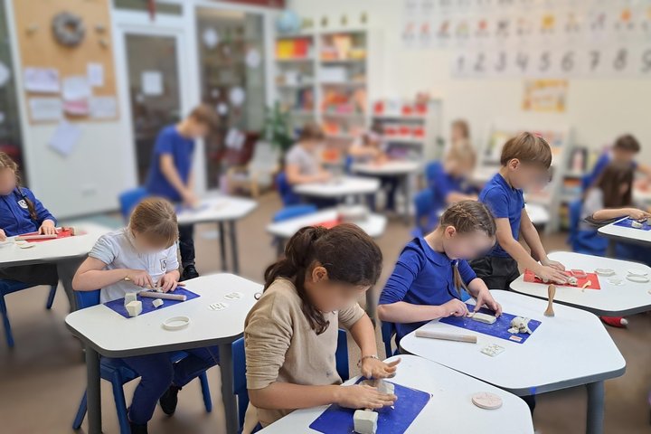 Nuotraukoje vaizduojama nemaža mokyklinė klasė. Joje prie suolų sėdi mokiniai, kurie iš balto molio lipdo įvairias figūras. Pro klasės langus matosi, kad lauke sutemę. Galima manyti, kad nuotauka daryta anskti ryte. Vaikai dėvi tamsiai mėlynas mokyklines