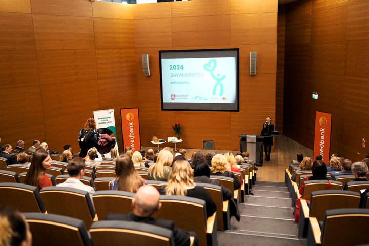 Matoma auditorija su žemyn besileidžiančiomis kėdžių eilėmis. Ant priekinės sienos kabo ekranas, kuriame skaidrė su užrašu "2024 savanorystės apdovanojimai". Scenoje stovi juodai apsirengusi moteris su mikrofonu. Prie abiejų sienų pastatyti Swedbank stend