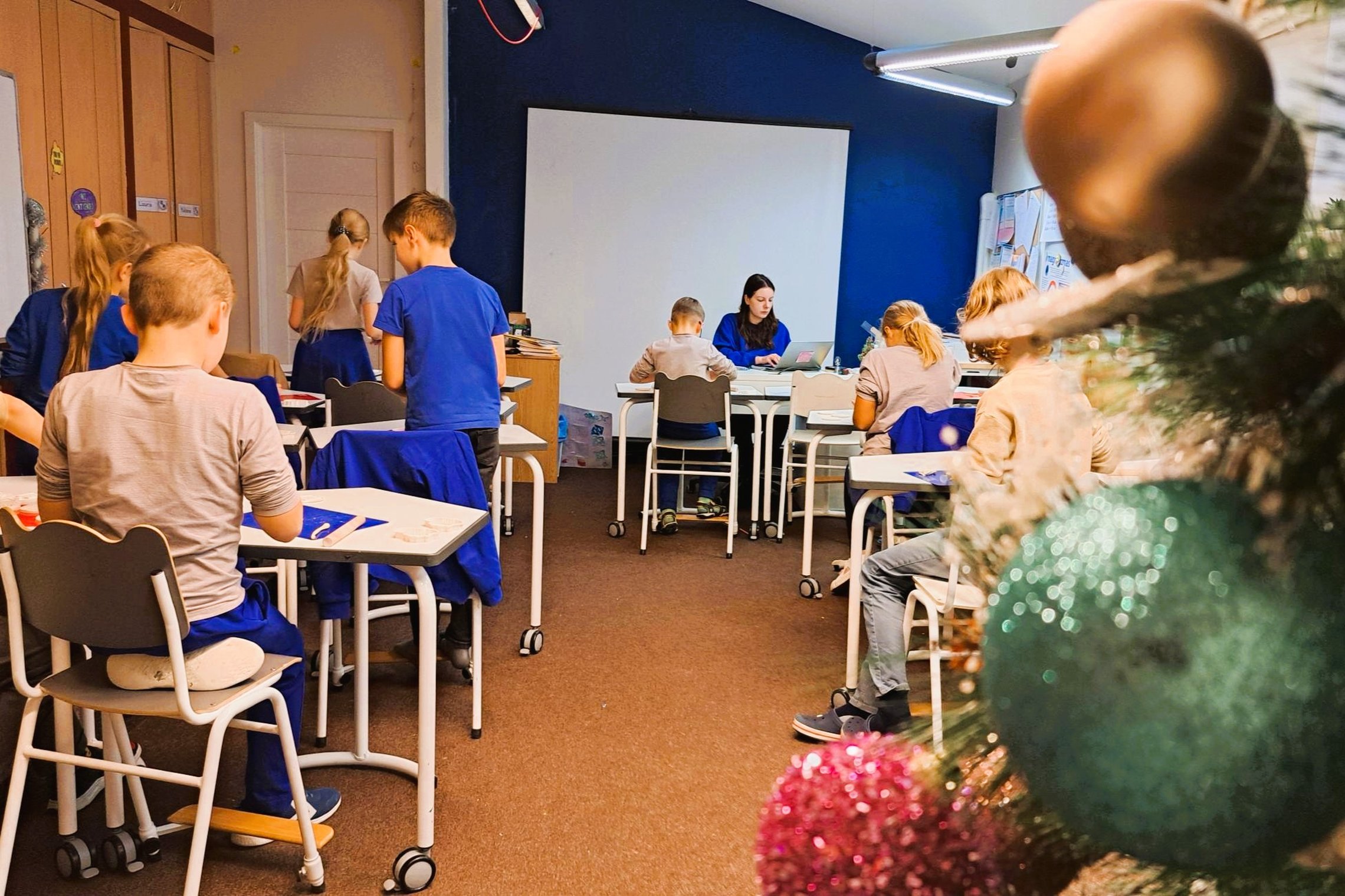 Nuotrauka padaryta iš klasės galo. Dešinėje pusėje iš arti matoma Kalėdų eglutė su daug žaisliukų. Vaikai stovi arba sėdi prie savo suolų, klasės priekyje prie mėlynos sienos su baltu ekranu sėdi ir kompiuteriu dirba moteris. Nesimato, ką vaikiai tiksliai