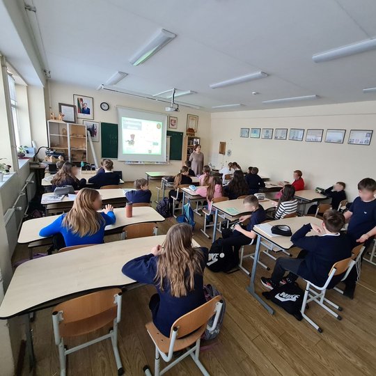 Vyksta istorijos pamoka. Mokiniai sėdi suoluose, mokytoja stovi klasės priekyje.