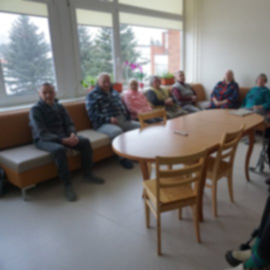 Širvintų rajono socialinių paslaugų centro lankytojai palei sienas susėdę kabinete. Kambario centre apvalus medinis stalas.