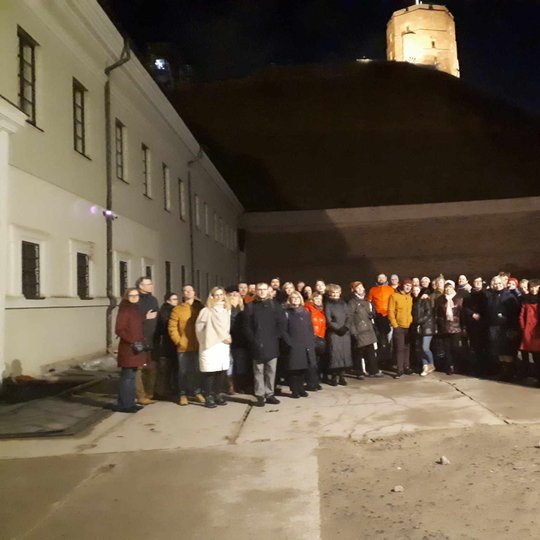 Ekskursantai Nacionalinio muziejaus kieme. Virš jų apšviestas Gedimino pilies bokštas.