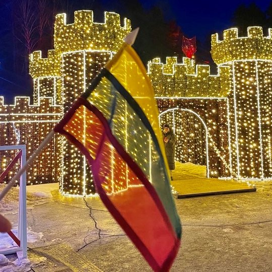 Trispalvės vėliavėlė, laikoma rankoje. Fone šviečia Lumina park pilies instaliacija.