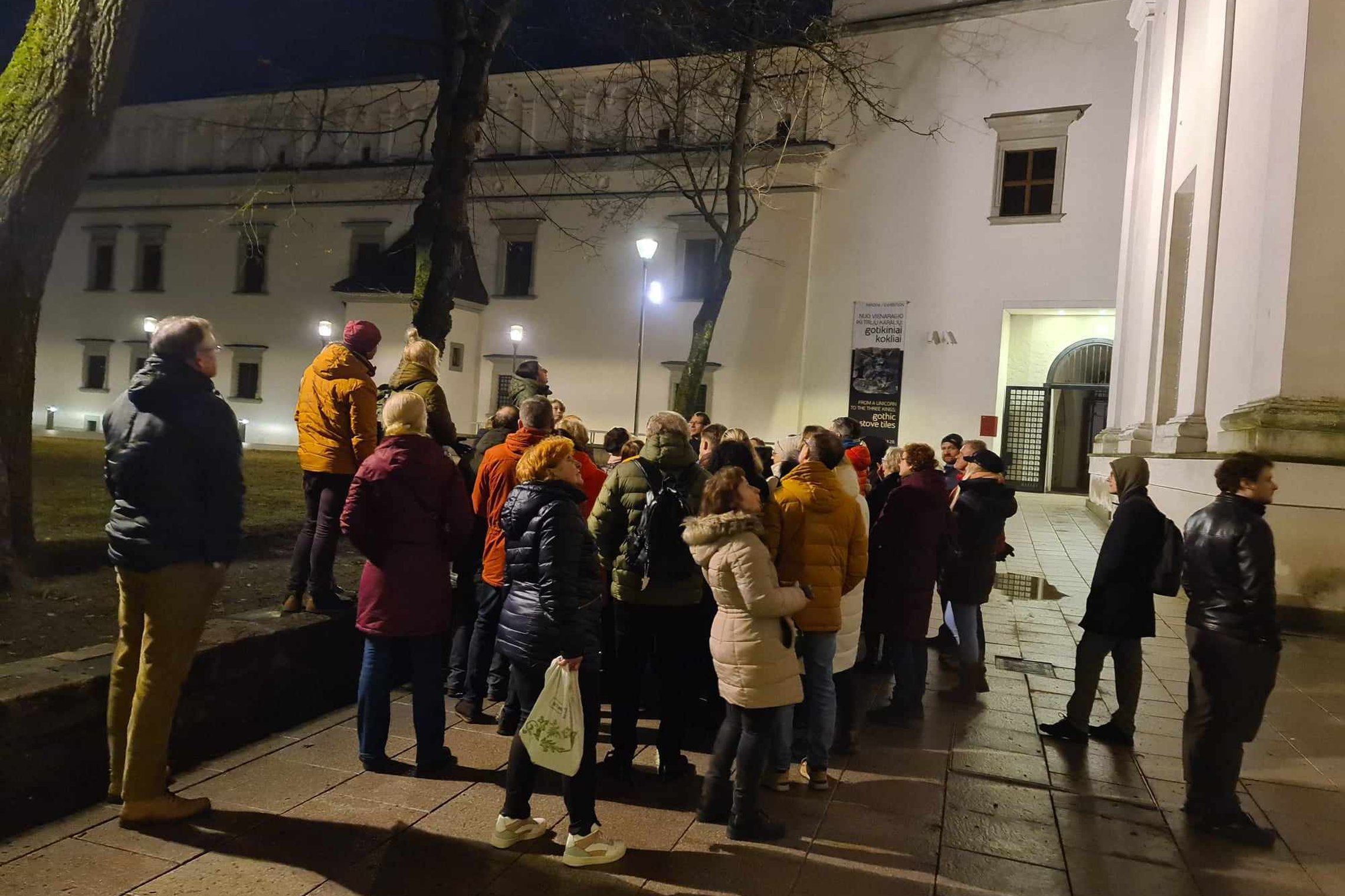 Grupė ekskursijos dalyvių Katedros aikštės šiaurinėje pusėje, aplink jau sutemę.