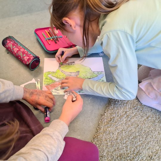 Dvi mergaitės ant klasės grindų spalvina Kreivosios pilies piešinį.