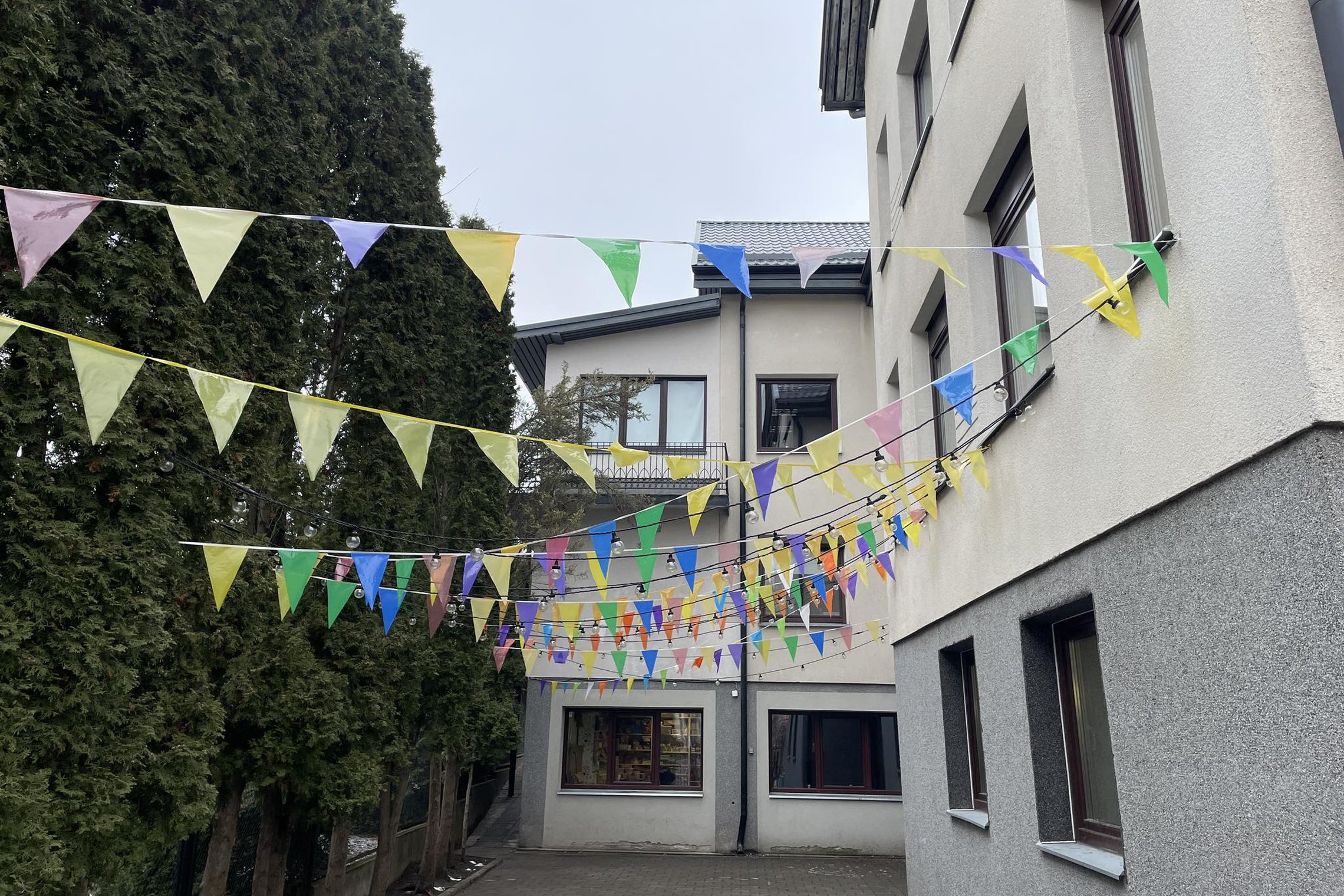 Matomas pastato kampas, įvažiavimas prie pastato ir krūmai. Tarp pastato ir krūmų pakabinta daug spalvingų vėliavėlių.