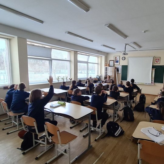 Klasėje vyksta pamoka. Klasės gale, suole sėdintis mokinys kelia ranką.
