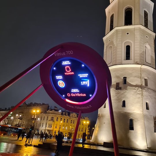 Pirmame plane šiek tiek kairiau yra raudona platforma ant kurios sumontuotas laikrodis, kuris skaičiuoja laiką, likusį iki septyni šimtojo Vilniaus miesto gimtadienio. Nuotraukos fone stūkso katedros varpinė.