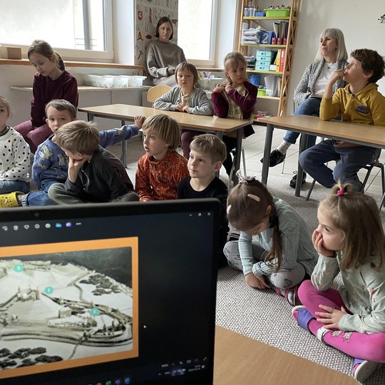 Vaikai susėdę ant žemės klausosi pasakojimo apie Vilniaus pilis.