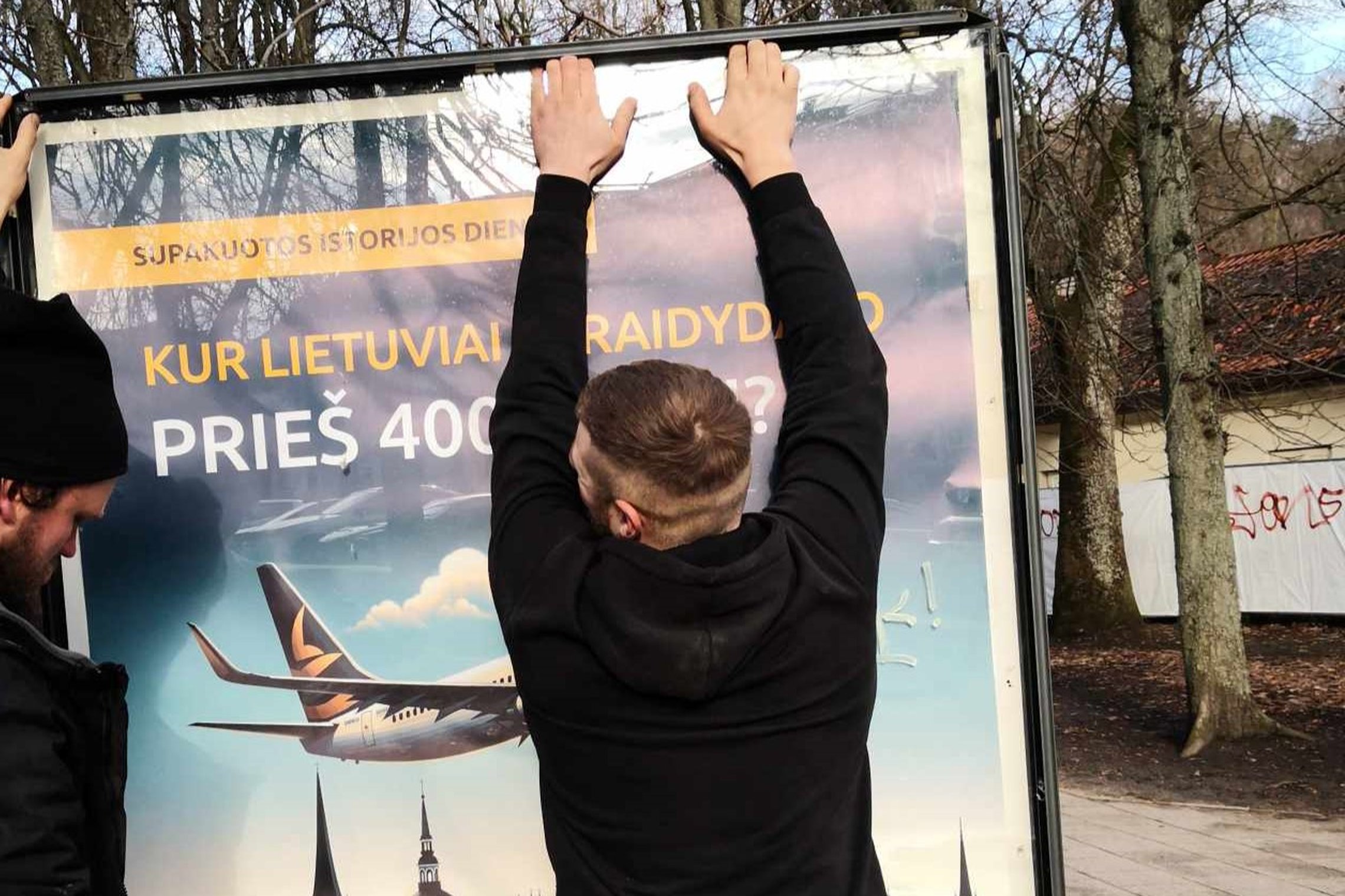 Du vyrai keičia informacinio stendo plakatą, ant kurio vaizduojamas lėktuvas, matosi klausimas: Kur lietuviai skraidydavo prieš keturis šimtus metų?