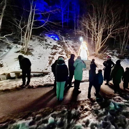 Matosi žmonės Kalnų parko take, kuris padengtas sniegu. Sniege švyti šviesų instaliacijos.